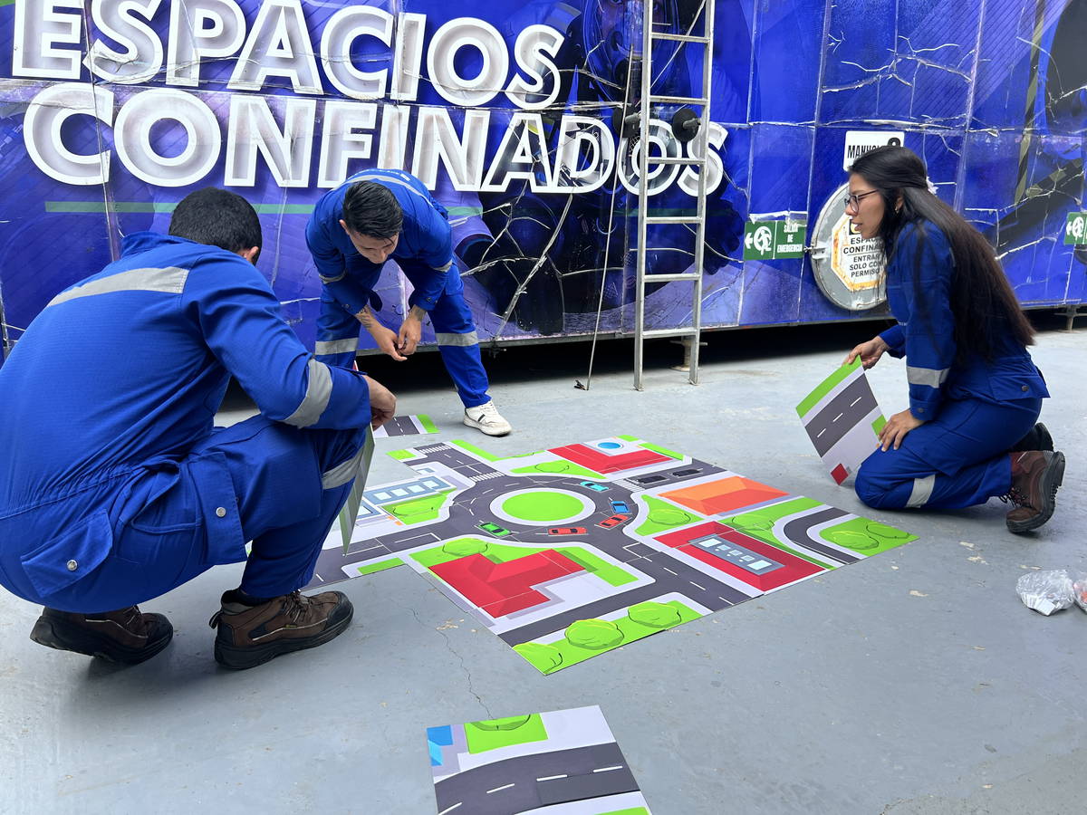 Trabajadores en overol azul resolviendo un juego de tablero de gran formato desplegado en el piso, frente a un pendón de espacios confinados.