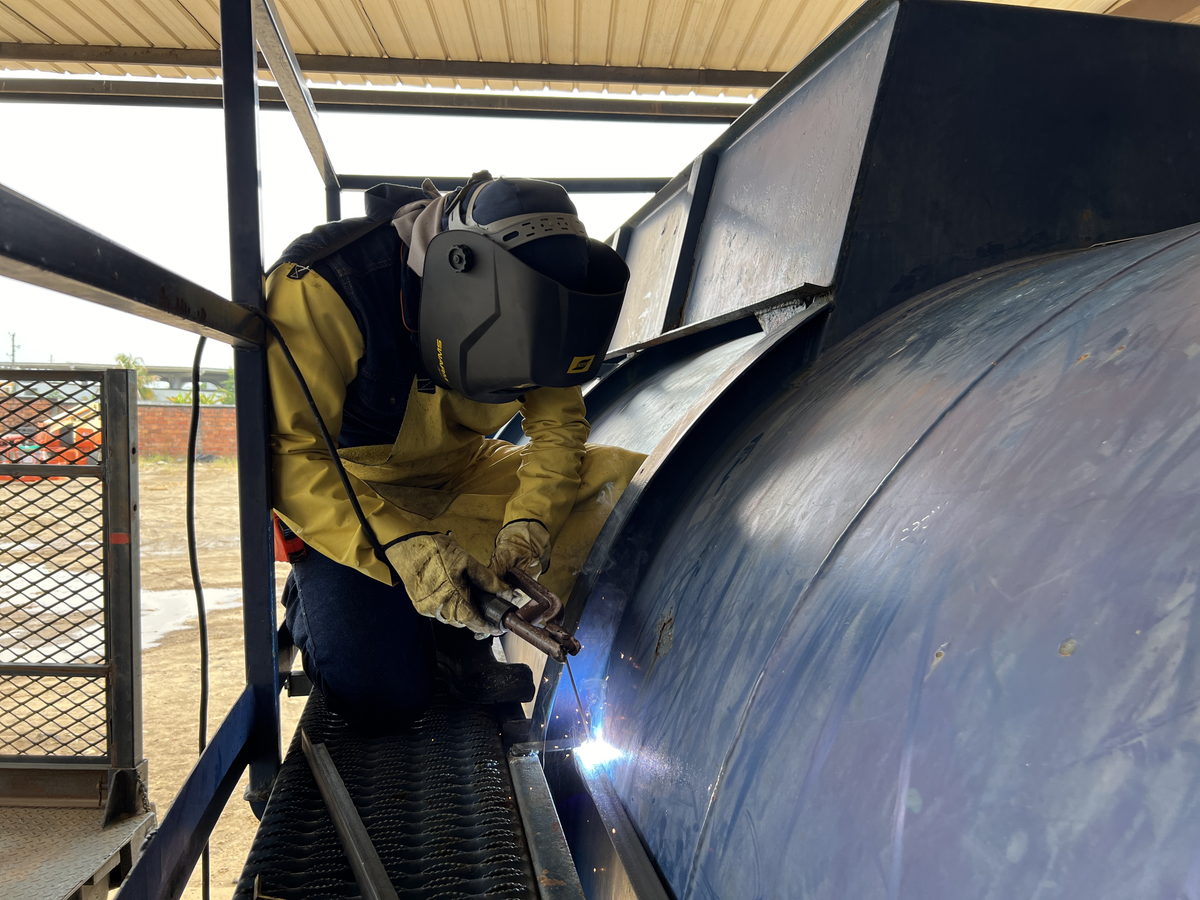 Soldador con careta y guantes de cuero trabajando sobre la superficie curva de un tanque metálico.