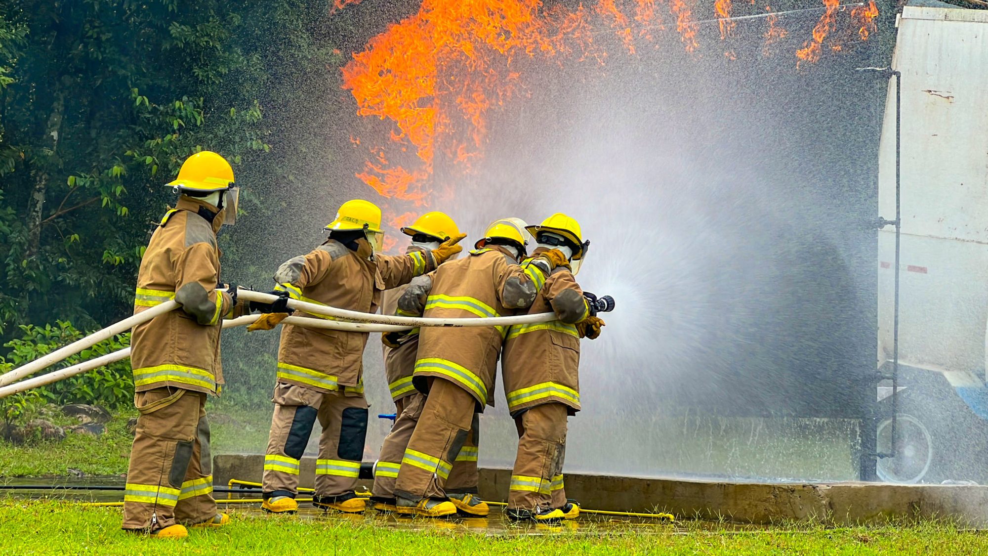 Brigada de emergencias en entrenamiento de control y extinción de fuego, avanzando con línea de manguera frente a un incendio controlado.